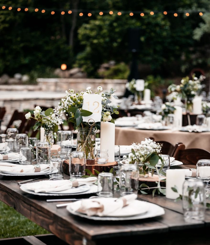 Outdoor reception with wooden dining tables set with place settings, flowers, and candles.